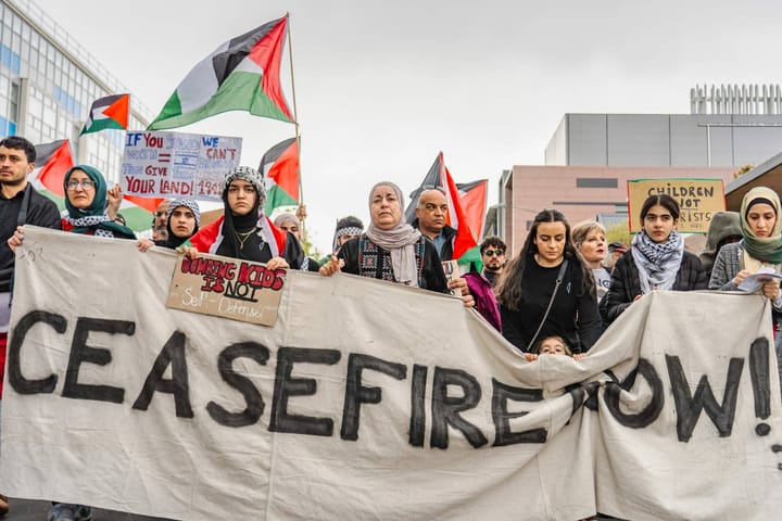 A line of mostly women, some in hijab, carrying a banner that says "CEASEFIRE NOW!" 